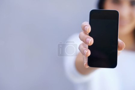 Foto de Tienes que coger este teléfono. Retrato de una joven de pie sobre un fondo gris y sosteniendo su teléfono móvil - Imagen libre de derechos