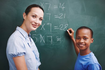 Photo for Patient and caring teacher. A young boy and his teacher looking at each other while he does sums on the board - Royalty Free Image