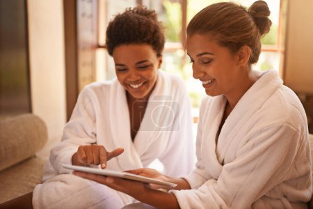 Foto de Vamos a empezar esta terapia... dos amigos en albornoces usando una tableta digital en un spa - Imagen libre de derechos