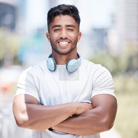 Photo for Fitness man wearing headphones around his neck while looking at the camera and smiling. Cheerful male runner standing with his arms crossed while out in the city for a workout. - Royalty Free Image