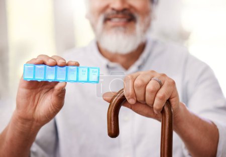 Photo for This helps me to remember what medication to take. a senior man sitting with a walking stick and holding up a weekly pill box - Royalty Free Image
