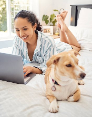Téléchargez les photos : Refroidir avec mon chien. une jeune femme utilisant un ordinateur portable tout en se relaxant sur son lit à la maison - en image libre de droit