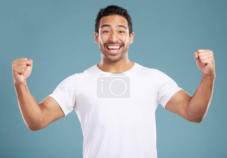 Photo for Handsome young mixed race man celebrating victory or success while standing in studio isolated against a blue background. Hispanic male cheering and pumping his fists at success or achievement. - Royalty Free Image
