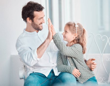Téléchargez les photos : Joyeux caucasien jeune père donnant à sa petite fille une haute cinq pour soutenir et motiver tout en étant assis sur une chaise ensemble à la maison. Homme joignant la main à son enfant profitant d'une journée ensemble. - en image libre de droit