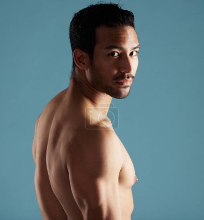 Foto de Handsome young hispanic man standing shirtless in studio isolated against a blue background. Mixed race topless male athlete looking confident, healthy and fit. Exercising to increase his strength. - Imagen libre de derechos