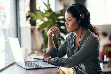 Foto de No es difícil ver por qué el freelance es tan tentador. una mujer joven usando un ordenador portátil y auriculares en un café - Imagen libre de derechos