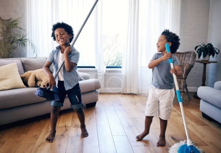 Photo for Two mixed race little boys playing with a mop and broom in the lounge at home. Excited siblings having fun playing with cleaning supplies at home. children singing and playing air guitar. - Royalty Free Image