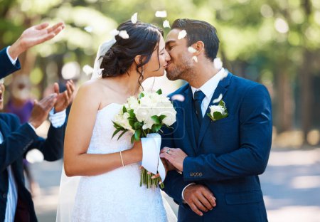 Téléchargez les photos : Heureux mariés partageant un baiser entouré d'invités et se douchant de pétales de rose confettis après la cérémonie de mariage. - en image libre de droit