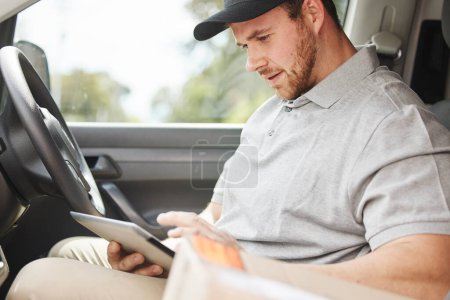 Foto de ¿Cuál es la dirección de nuevo. un joven y guapo repartidor usando una tableta mientras está sentado en su furgoneta - Imagen libre de derechos