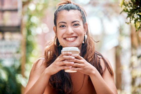 Téléchargez les photos : Joyeux jeune femme métissée profitant d'une tasse de café lors d'une pause dans un café de la ville. Une femme ne boit qu'une boisson chaude dans une tasse de papier à emporter jetable tout en se relaxant devant un restaurant. - en image libre de droit
