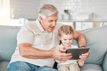 Foto de Un niño necesita un abuelo. una adorable niña usando una tableta digital mientras está sentada en casa con su abuelo - Imagen libre de derechos