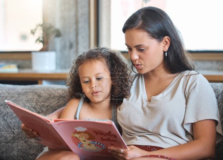 Foto de Amar a la madre hispana y a su hijita sentadas en casa y leyendo un libro de cuentos juntas. Madre enseñando a la niña a leer mientras está sentada en el sofá en casa. - Imagen libre de derechos