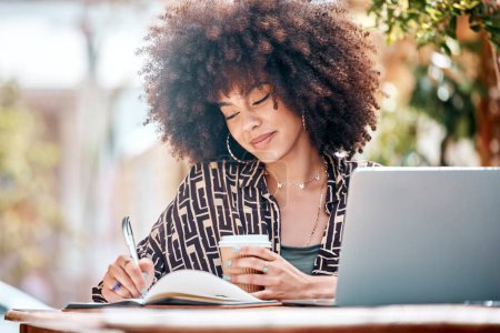 Photo for Young happy stylish mixed race businesswoman with a curly afro writing in a diary drinking coffee and working on a laptop sitting outside at a cafe. Hispanic female student studying at a restaurant. - Royalty Free Image