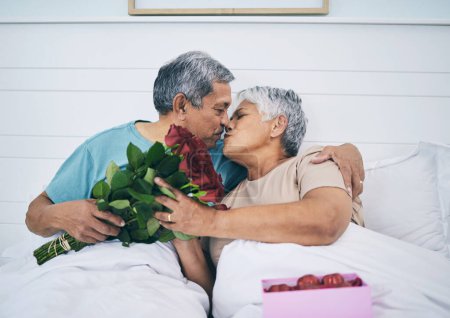 Photo for Love, elderly couple and kiss with roses in bed, romance and affection in home. Flowers, senior man and woman in bedroom for intimacy, care and enjoying quality time together with floral bouquet gift. - Royalty Free Image
