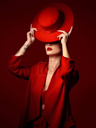 Photo for Hat, fashion and a model woman on a red studio background for elegant or trendy style. Aesthetic, art or confidence with a young female person in an edgy and classy outfit, suit or clothes for beauty. - Royalty Free Image