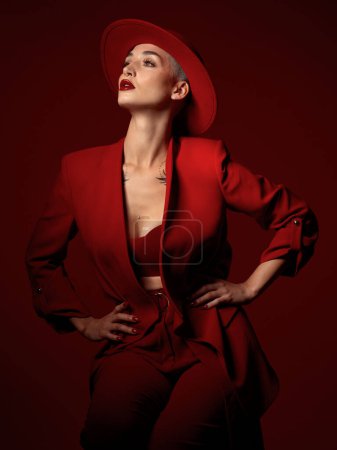 Photo for Confident, fashion and a model woman on a red studio background for elegant or trendy style. Aesthetic, art and beauty with a young female person standing hands on hips in an edgy or classy suit. - Royalty Free Image