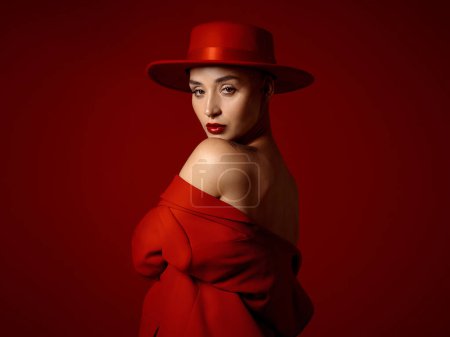 Photo for Portrait, fashion and shoulder with a woman on a red studio background for elegant or trendy style. Aesthetic beauty, art and hat with a young female model looking edgy or classy in a unique outfit. - Royalty Free Image