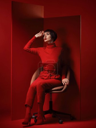 Photo for Thinking, fashion and a model woman on red background in studio for elegant, chic or trendy style. Aesthetic, art and beauty with an edgy person in unique clothes suit, makeup and cosmetics on chair. - Royalty Free Image