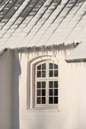 Téléchargez les photos : Bâtiment vide avec barrage de glace sur le toit par une froide journée d'hiver. L'extérieur d'une maison ou d'une maison avec de la neige sur le toit par un après-midi ensoleillé. Froid sur une église historique dans un village. - en image libre de droit