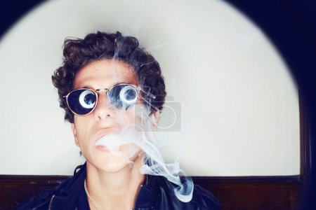 Téléchargez les photos : Portrait de l'homme fumant, lunettes de soleil et mode grunge avec attitude rockstar sur fond blanc sous les projecteurs. Style punk cool, créatif et fumé, visage confiant du beau mannequin masculin en studio - en image libre de droit