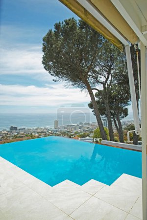 Photo for Scenic view of infinity swimming pool overlooking city and ocean in background. Luxury outdoor blue pool on a tiled deck, patio and veranda of a condo, home or hotel. Cityscape and skyline with sea. - Royalty Free Image