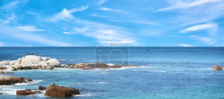 Téléchargez les photos : Rochers à une plage sur un fond bleu ciel nuageux avec espace de copie. Océan calme et majestueux à l'horizon et côte rocheuse. Paysage pittoresque de la mer pour des vacances d'été ou une escapade. - en image libre de droit