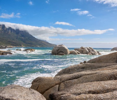 Photo for A calming ocean view of the rocky coast of Camps Bay in Cape Town, South Africa. Boulders at a beach with the tide and current coming in and Table Mountain on the horizon. Tidal waves washing ashore. - Royalty Free Image