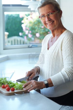Foto de Retrato de la mujer mayor en la tabla de cortar, verduras y cuchillo en la cocina de alimentos para cocinar. Persona feliz en vasos cortando plantas en mostrador, lechuga y tomates, nutrición y dieta saludable en el hogar. - Imagen libre de derechos