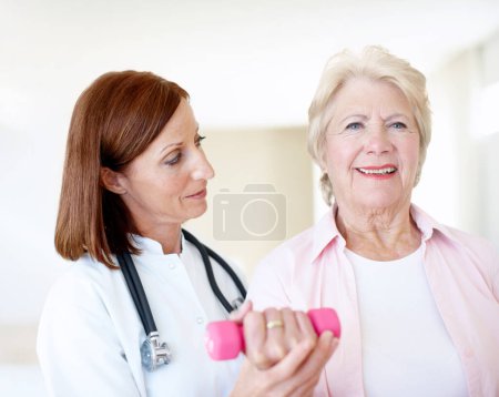 Photo for Buoyed by her progress. Determined elderly female is assisted by her nurse as she lifts a dumbbell - Royalty Free Image