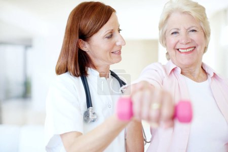 Photo for The strength of her resolve - Senior Care. Smiling elderly female is assisted by her nurse as she lifts a dumbbell - Focus on background - Royalty Free Image