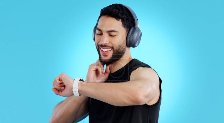 Photo for Happy man, headphones and checking watch for pulse in fitness against a blue studio background. Male person, athlete or model looking at wristwatch for monitoring performance or heart rate on mockup. - Royalty Free Image
