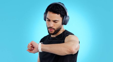 Photo for Man, headphones and checking watch in fitness for performance against a blue studio background. Male person, athlete or model looking at wristwatch for monitoring or tracking steps on mockup space. - Royalty Free Image