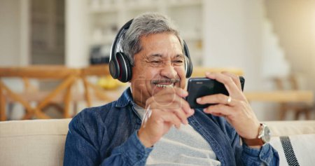 Photo for Senior, man and headphone with phone in living room for streaming, entertainment and mobile app. Elderly person, smile and retirement with technology with laugh for funny video, movie or film in home. - Royalty Free Image