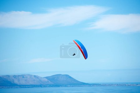 Téléchargez les photos : Pilote, parachute et parapente dans le ciel bleu pour le vol, la liberté et le courage avec un sport extrême. Athlète, glisse et sans peur en plein air fitness pour l'aventure, la santé et les sports par l'océan avec la montagne. - en image libre de droit