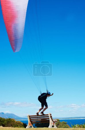 Photo for Man, parachute or paragliding sport in launch exercise, healthy adventure or extreme fitness for wellness. Person, preparation and fearless by bench for flight with helmet and safety gear by blue sky. - Royalty Free Image