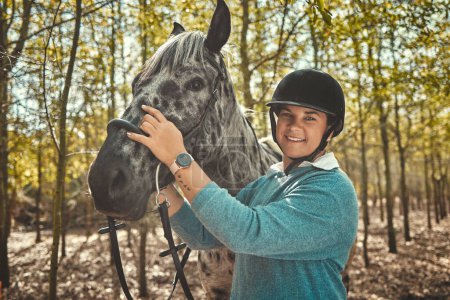 Téléchargez les photos : Portrait de femme à cheval en forêt, sourire et fierté pour la compétition, course ou dressage avec des arbres. Sport équestre, visage de jockey ou cavalier avec animal dans les bois pour l'aventure, l'entraînement et les soins - en image libre de droit