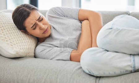 Woman, stomach pain and stress on sofa, sick or menstruation with anxiety, gas or virus in home living room. Girl, abdomen and emergency for gut health, constipation or cramp on lounge couch in house.