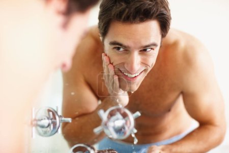 Photo for Man, washing face and smile with water for cleaning in bathroom, mirror and reflection for bacteria and facial hygiene. Person, hand and sink for skincare with soap for grooming, health or wellness. - Royalty Free Image