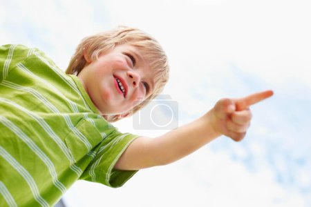 Foto de Niño, niño feliz y apuntando al cielo azul al aire libre en ángulo bajo, maqueta de verano y mostrar dirección. Joven lindo niño, dedo y mano gesto en las nubes, jugando y la libertad para la educación de abajo con sonrisa. - Imagen libre de derechos