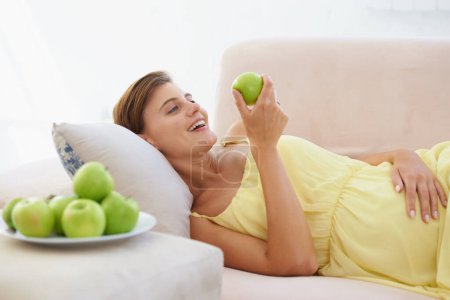 Foto de Mujer embarazada, manzana y sonrisa en sofá para la salud, nutrición y bienestar en la sala de estar del apartamento. Persona, fruta y feliz en el sofá del salón para relajarse, embarazo y dieta saludable con vitaminas. - Imagen libre de derechos