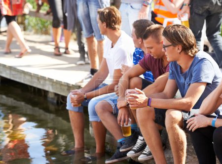 Photo for Drink, beer and people on holiday at lake, water or deck in summer at music festival with men. Friends, party and relax at dam with alcohol, conversation and fun at social, event or sitting on pier. - Royalty Free Image