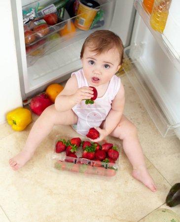 Photo for Toddler, little girl and strawberry for eating in kitchen with open, fridge and door with above view. Youth, kid and hunger for fresh, organic and fruit for healthy, balanced or snack for development. - Royalty Free Image