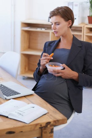 Foto de Mujer de negocios, embarazada y comiendo zanahorias para una dieta saludable, nutrición o comida en la oficina. Mujer o empleada en embarazo o maternidad con portátil y verduras orgánicas naturales en el lugar de trabajo. - Imagen libre de derechos