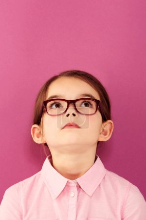 Photo for Glasses, child and looking up with thinking geek and modern fashion in a studio with serious face. Kid, eyewear and sight with outfit and nerd with accessory and pink background with confidence. - Royalty Free Image
