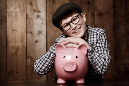 Téléchargez les photos : Piggy Bank, portrait et garçon enfant sourire avec l'épargne, la croissance ou le succès d'investissement dans sa maison. Finance, apprentissage et visage de l'enfant excité avec boîte à argent pour l'éducation future, le financement ou la sécurité. - en image libre de droit