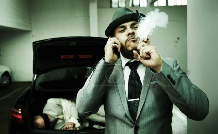 Photo for Phone call, car and mafia with hostage in trunk for negotiation, kidnapping danger and crime. Smoking, gangster criminal and business person in boot for abduction, terrorism and money in parking lot. - Royalty Free Image