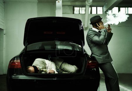 Photo for Phone call, smoking and criminal with hostage in trunk for negotiation, kidnapping ransom and crime. Mafia, gangster and business person in boot for abduction, robbery and money in parking lot. - Royalty Free Image