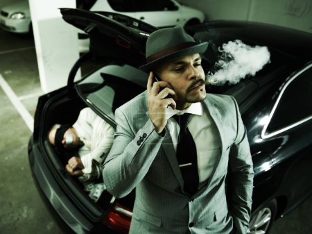 Photo for Phone call, car and man with abduction in trunk for negotiation, kidnapping ransom and crime. Mafia, gangster criminal and person smoking in boot for terrorism, danger and robbery in parking lot. - Royalty Free Image