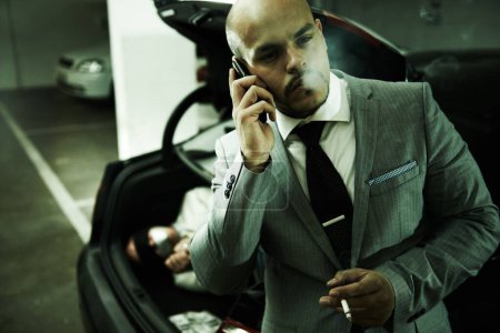 Photo for Mafia, car and man with hostage in trunk for negotiation, kidnapping ransom or crime. Phone call, gangster criminal and business person smoking in boot for abduction, danger or robbery in parking lot. - Royalty Free Image
