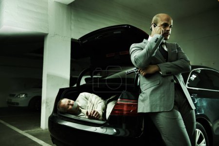 Photo for Phone call, ransom and man with hostage in car for negotiation, kidnapping danger and crime. Mafia, gangster criminal and business person in boot for robbery, abduction and terrorism in parking lot. - Royalty Free Image